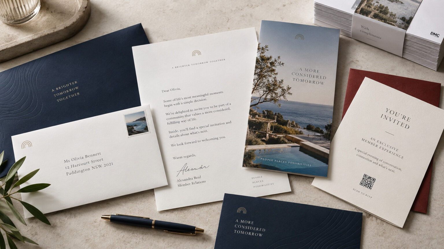 Elegant invitation suite on a beige surface: navy card, white letter, photo brochure, and a fountain pen.
